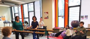 A group of women exercise in a room together, smiling at the camera.